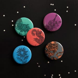 May include: Five round buttons with unique illustrations. The buttons feature a teal owl, a red dragon and wolf, a blue floral design, a purple abstract design, and a gray and orange mask design. The buttons are on a black background with star-shaped confetti.