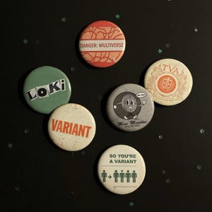 May include: A collection of six circular pinback buttons with various designs and text. The buttons feature phrases like "DANGER: MULTIVERSE," "LOKI," and "VARIANT." The color palette includes red, green, white, and gray.