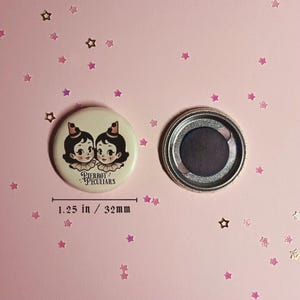 May include: A round button magnet measuring 1.25 inches in diameter. The front features an illustration of two figures with dark hair, ruffled collars, and small hats. The text Pierrot Peculiars is printed below. The back shows a circular magnet.