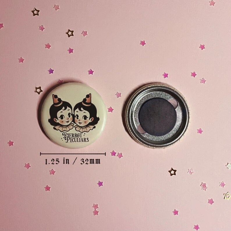 May include: A round button magnet measuring 1.25 inches in diameter. The front features an illustration of two figures with dark hair, ruffled collars, and small hats. The text Pierrot Peculiars is printed below. The back shows a circular magnet.