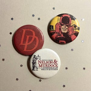 May include: Three circular pin-back buttons. One is dark red with a red DD logo. Another is yellow and black featuring a comic book character in a red mask. The third is white with a black illustration of Lady Justice and the text Nelson & Murdock.
