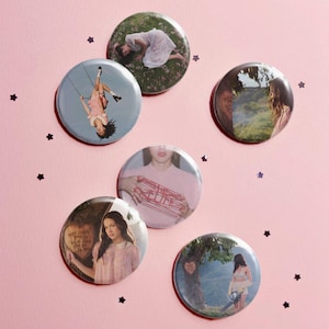 May include: A collection of six circular pin-back buttons featuring various photographic prints. The images include a person on a swing, a person in a field, a person with red string, and a person holding a heart-shaped sign with text.
