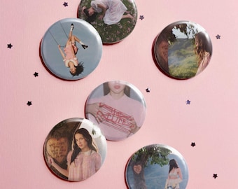 May include: A set of six circular pin-back buttons featuring various photographic prints. The images include a person lying in grass, a person on a swing, a landscape, a person holding red string, and a person standing near a tree.