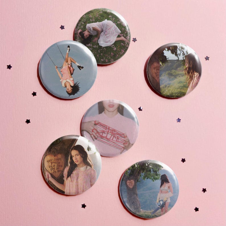 May include: A set of six circular pin-back buttons featuring various photographic prints. The images include a person lying in grass, a person on a swing, a landscape, a person holding red string, and a person standing near a tree.