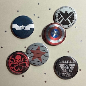 May include: A collection of six round pinback buttons featuring various designs. The buttons showcase imagery from the Marvel universe, including Captain America's shield, the Winter Soldier's star, and S.H.I.E.L.D. logos. The color palette includes blue, red, and silver.