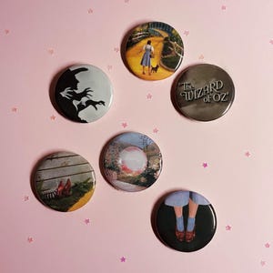 May include: A collection of six round pinback buttons featuring imagery from The Wizard of Oz. The buttons display scenes and characters, including Dorothy and Toto on the yellow brick road, the Wicked Witch, the title, and the ruby slippers.