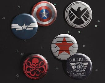 Op de afbeelding: Een set van zes ronde buttons met diverse grafische ontwerpen: een rood, wit en blauw schild met een ster, een zwart-wit adelaarembleem, een donkerblauwe button met een witte ster, een rode ster op een witte achtergrond, een rode schedel met tentakels en een zwarte button met de tekst S.H.I.E.L.D. ACADEMY SCIENCE AND TECHNOLOGY DIVISION.