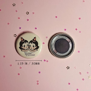 May include: A round button magnet measuring 1.25 inches in diameter. The front features an illustration of two figures with dark hair, ruffled collars, and small hats. The text Pierrot Peculiars is printed below. The back shows a circular magnet.