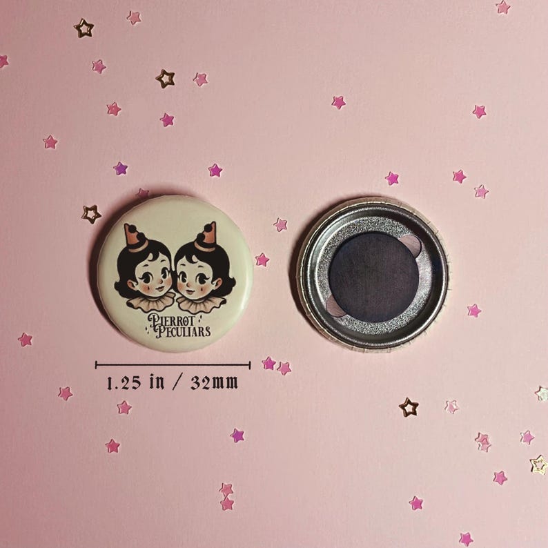 May include: A round button magnet measuring 1.25 inches in diameter. The front features an illustration of two figures with dark hair, ruffled collars, and small hats. The text Pierrot Peculiars is printed below. The back shows a circular magnet.