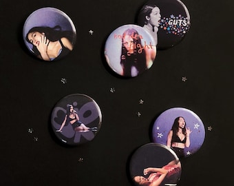 May include: Five circular pin-back buttons featuring photographic prints of a person. One button displays the text GUTS with colorful stars, another says bad idea right, and the others show various poses. Small silver star-shaped confetti are scattered around.