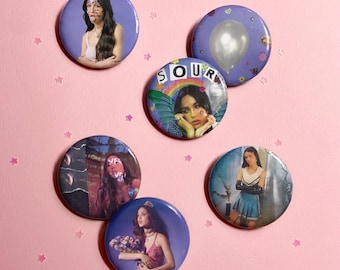 May include: Five circular pin-back buttons featuring Olivia Rodrigo. One shows her with face stickers, one has a balloon, one displays the text SOUR with her wearing wings, one shows her in a dark setting, and one shows her in a cheerleader outfit.