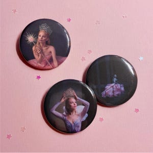 May include: Three circular pin-back buttons featuring photographic prints. One shows a person in a crown holding a wand, another shows a person in a crown with arms raised, and the third shows a person in a pink gown standing on a staircase.