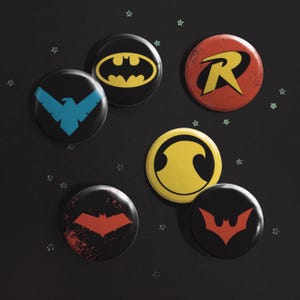 May include: A set of six circular pin-back buttons featuring various superhero-themed emblems. The designs include a yellow Batman bat symbol, a red Robin R, a blue Nightwing bird, a yellow circle, a red bat symbol, and a red bat emblem.