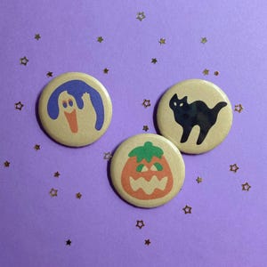 May include: Three round Halloween-themed buttons. One button features a blue ghost with an orange beak, another a black cat, and the last an orange jack-o'-lantern with a green stem. The buttons are on a purple background with gold star confetti.