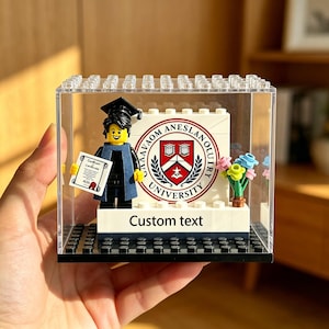 Puede incluir: Una escena de graduación de Lego en una vitrina transparente. La escena incluye una figura de Lego con birrete y toga de graduación sosteniendo un diploma. El fondo presenta un logotipo universitario y las palabras "Custom text".