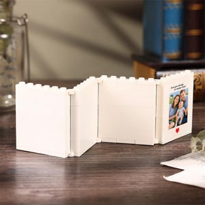 May include: A white, foldable, multi-panel picture frame constructed from interlocking building blocks. The frame features a hinged design with three sections, one of which displays a photograph of a family with the text, Love you always, no matter what, and a small red heart icon.