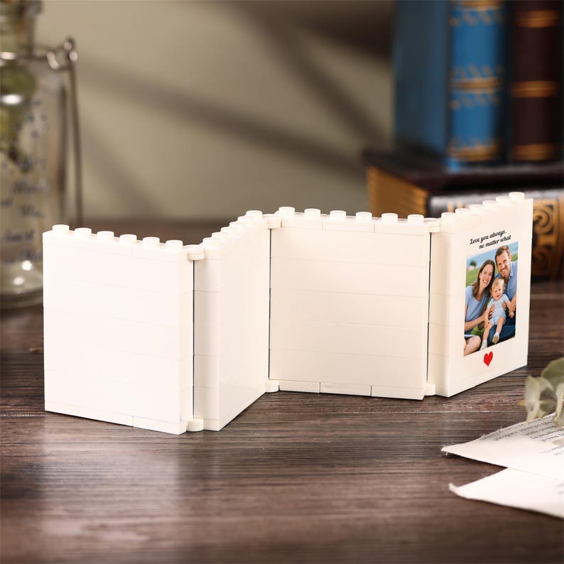 May include: A white, foldable, multi-panel picture frame constructed from interlocking building blocks. The frame features a hinged design with three sections, one of which displays a photograph of a family with the text, Love you always, no matter what, and a small red heart icon.