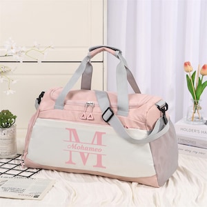 Peut inclure: Un sac de sport rose et blanc avec des sangles et des détails gris. Le sac porte le nom "Mohameo" et la lettre "M" en rose. Il possède une poche latérale en filet et une bandoulière amovible. Idéal pour le voyage ou la salle de sport.
