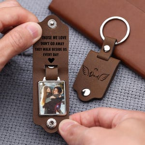 May include: Brown leather keychain with a silver-toned photo frame. The keychain has the text "THOSE WE LOVE DON'T GO AWAY THEY WALK BESIDE US EVERY DAY" and a heart symbol. A second keychain has the word "Dad" with angel wings.