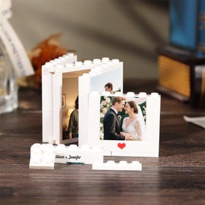 May include: A white plastic building block photo frame set featuring two vertical frames and a base piece. The frames display photographs of a couple and a person. The base piece is printed with the text William & Jennifer and a small red heart.