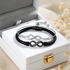 May include: Two infinity symbol bracelets. One is black leather with a black infinity charm, the other is silver with a silver infinity charm. The black bracelet has the names "Mireyal" and "Ericayl" engraved. The silver bracelet has the names "Name 1" and "Name 2" engraved.