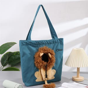 May include: Teal tote bag with a lion design. The bag features a plush lion's mane and face, with the name "Chloé" printed on the side. The bag has long handles for carrying.