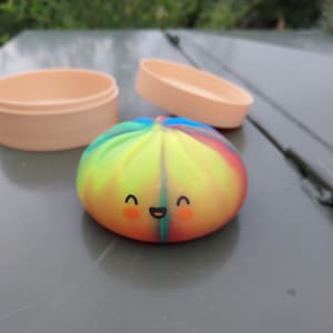May include: A round, multi-colored tie-dye silicone dumpling toy featuring a printed smiling face with orange cheeks. The toy is accompanied by two separate, light peach-colored circular containers with matching lids, all resting together.