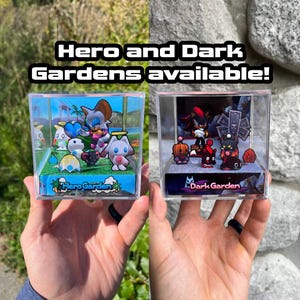 Könnte beinhalten: Zwei transparente Display-Würfel, jeweils mit Miniaturfiguren. Ein Würfel zeigt eine blau-grüne Landschaft mit Charakteren, beschriftet mit "Hero Garden". Der andere, "Dark Garden", zeigt eine dunklere Szene mit roten und schwarzen Figuren. Text: "Hero and Dark Gardens available!"