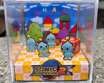 以下が含まれることがあります： ソニックアドベンチャー2のキャラクターが入った透明なプラスチックケース。ゲームのロゴが入った市松模様のプラットフォームに、4体の青いチャオのフィギュアが入っています。背景には「CHAOS」の文字とカラフルな風景が描かれています。