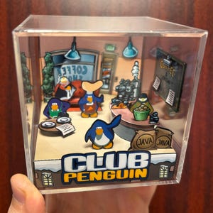May include: A clear acrylic display box featuring a Club Penguin diorama. The scene includes penguin characters in a coffee shop setting, with the words "Club Penguin" prominently displayed. The box is held by a hand.