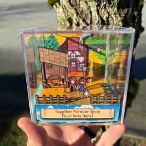 Könnte beinhalten: Ein Diorama in einer transparenten Acrylbox mit einer Miniaturszene mit zwei Figuren, einem Haus und dem Text "Together Forever Since [Your Date Here]" im Cartoon-Stil, mit einem Zaun und Bäumen.