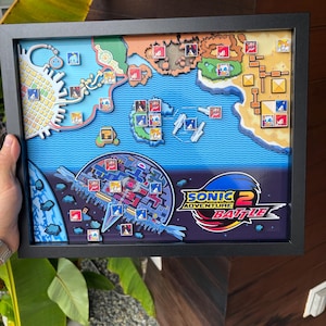 May include: A framed 3D shadow box display featuring a stylised map from the video game Sonic Adventure 2 Battle. The artwork includes layered terrain, water, and various character icons placed across the map, with the game logo in the corner.