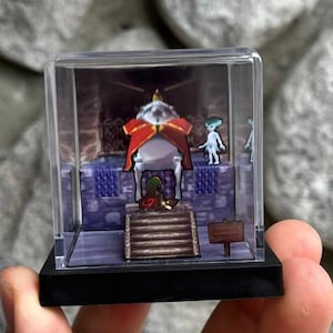 May include: A miniature diorama in a clear plastic case. The scene shows a character on a throne with two other figures. The throne room has a blue and purple colour scheme. A small sign is visible.