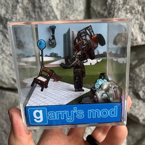 May include: A clear acrylic cube featuring a miniature diorama scene from Garry's Mod. The scene includes a character, a vehicle, and other game elements. The cube has the Garry's Mod logo in blue and white.