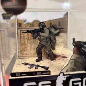 Puede incluir: Diorama en miniatura que representa una escena de videojuego. Dos soldados con equipo t&aacute;ctico apuntan con rifles. La escena incluye una caja de madera, un arma y el logotipo del juego. El fondo muestra un entorno des&eacute;rtico.