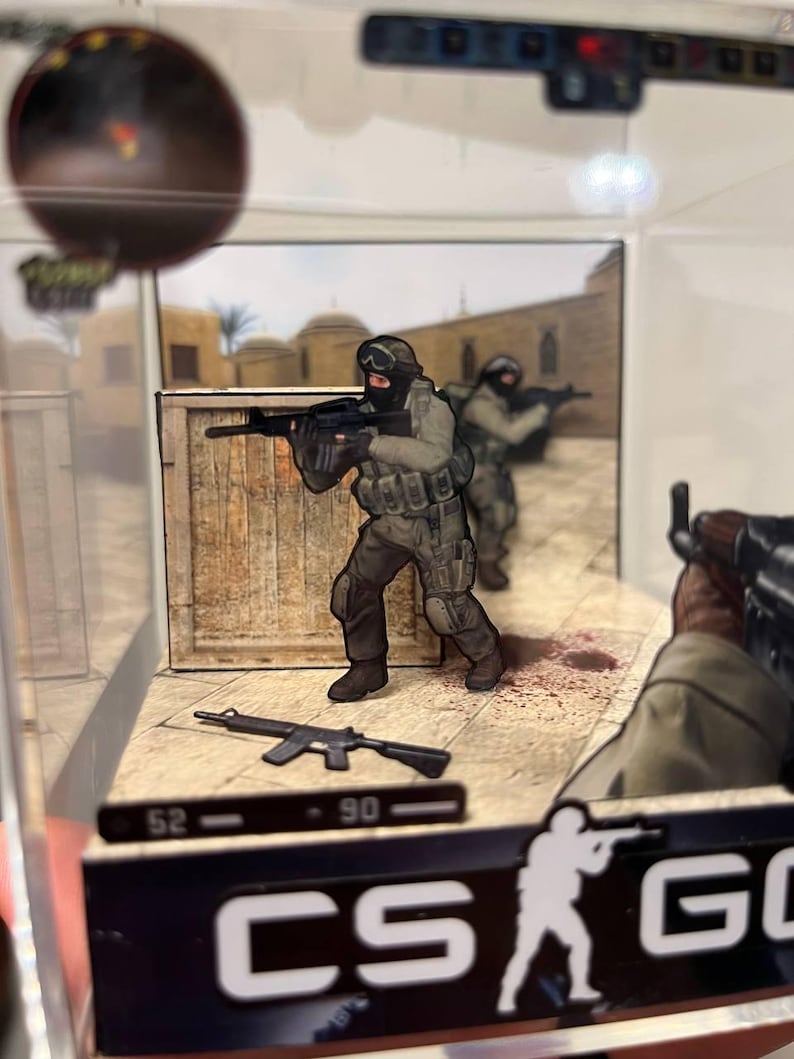 Puede incluir: Diorama en miniatura que representa una escena de videojuego. Dos soldados con equipo t&aacute;ctico apuntan con rifles. La escena incluye una caja de madera, un arma y el logotipo del juego. El fondo muestra un entorno des&eacute;rtico.