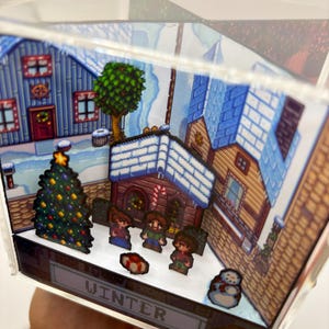 May include: A miniature winter village scene inside a clear acrylic cube. The scene features pixelated houses, a Christmas tree, and figures in the snow. The word "WINTER" is displayed on the base.