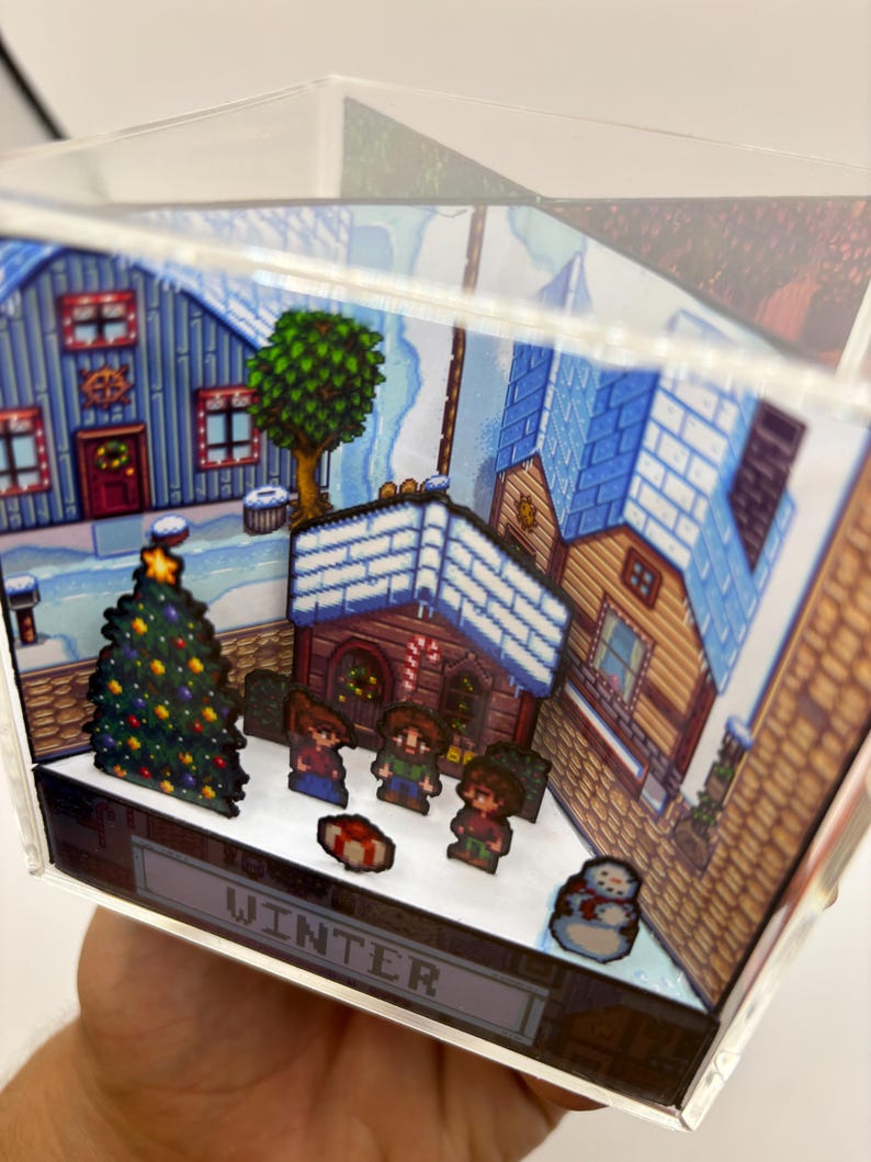 May include: A miniature winter village scene inside a clear acrylic cube. The scene features pixelated houses, a Christmas tree, and figures in the snow. The word "WINTER" is displayed on the base.