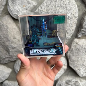 Puede incluir: Una vitrina de acrílico transparente con un diorama de Metal Gear Solid. La escena representa dos figuras en un entorno futurista, con el logotipo del juego en la parte inferior. El diorama se sostiene contra un fondo de piedras grises.