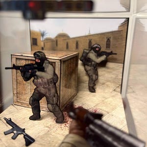 Puede incluir: Un diorama con figuras en miniatura de soldados con equipo de combate, sosteniendo rifles. La escena incluye una caja de madera, un fondo de edificio de estilo oriental y un efecto de salpicaduras de sangre en el suelo. Un rifle est&aacute; en el suelo.