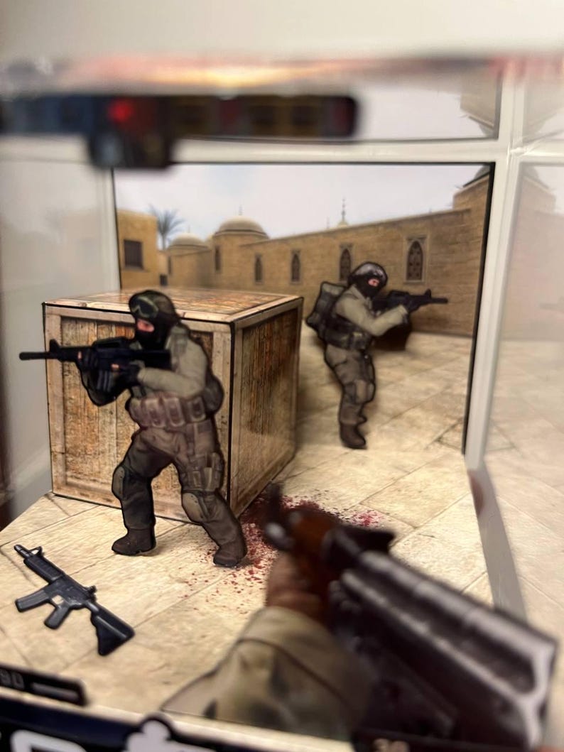 Puede incluir: Un diorama con figuras en miniatura de soldados con equipo de combate, sosteniendo rifles. La escena incluye una caja de madera, un fondo de edificio de estilo oriental y un efecto de salpicaduras de sangre en el suelo. Un rifle est&aacute; en el suelo.