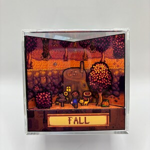 May include: A diorama featuring a pixelated autumn scene with a house, trees, and characters. The word "FALL" is displayed on a golden sign. The scene is enclosed in a clear, square frame.