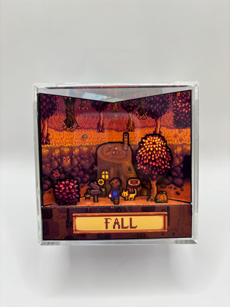May include: A diorama featuring a pixelated autumn scene with a house, trees, and characters. The word "FALL" is displayed on a golden sign. The scene is enclosed in a clear, square frame.