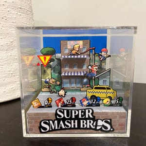 Könnte beinhalten: Ein Super Smash Bros. Diorama in einer transparenten Acrylbox. Die Szene zeigt verschiedene Charaktere in einer Stadtkulisse, mit dem Logo des Spiels am Boden. Das Diorama umfasst eine Ziegelfassade und ein gelbes Taxi.