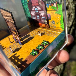 May include: A miniature diorama featuring a pixelated farm scene. The scene includes a house with a red roof, a wooden fence, and a field with flowers. The diorama is encased in a clear, rectangular box.