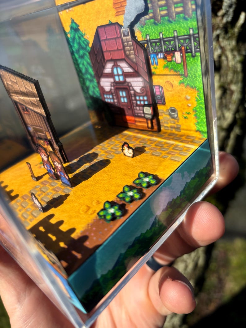 May include: A miniature diorama featuring a pixelated farm scene. The scene includes a house with a red roof, a wooden fence, and a field with flowers. The diorama is encased in a clear, rectangular box.