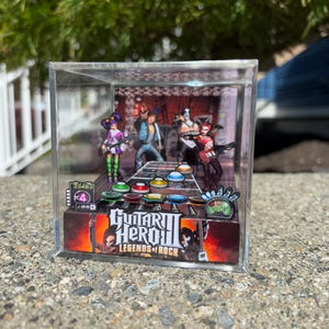 Op de afbeelding: Een miniatuur Guitar Hero II diorama in een heldere acryl kubus. De scène toont een band op het podium met een gitaarcontroller aan de onderkant. Het logo van het spel, "Guitar Hero II Legends of Rock", is zichtbaar.