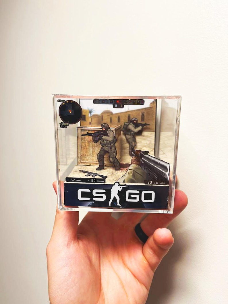 Puede incluir: Una vitrina de acr&iacute;lico transparente que presenta una escena del videojuego CS:GO. El diorama incluye figuras en miniatura de soldados, armas y elementos de la interfaz del juego. El logotipo de CS:GO se muestra prominentemente en la parte delantera.