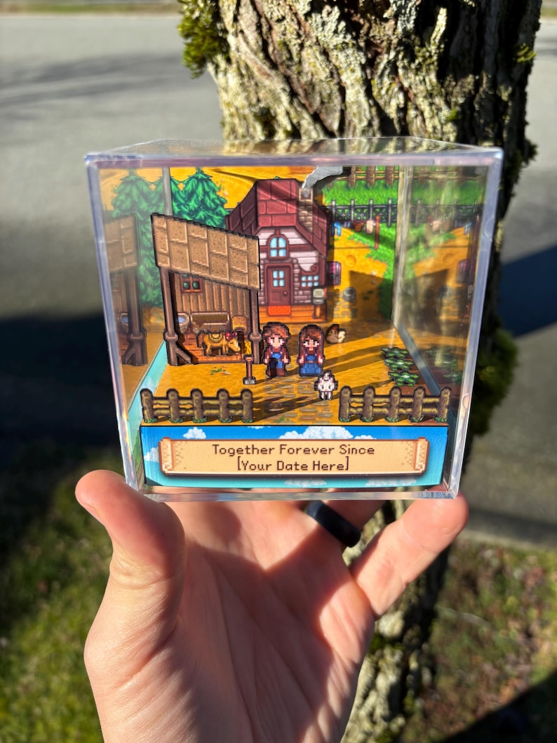 May include: A clear acrylic box diorama featuring a miniature scene with two figures, a house, and the text "Together Forever Since [Your Date Here]". The scene is set in a cartoonish style with a fence and trees.