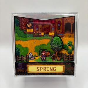May include: A diorama featuring a pixelated scene with a character, animals, and a building, all within a clear acrylic cube. The word "SPRING" is displayed on a golden banner at the bottom. The scene is set in a vibrant, colorful environment.